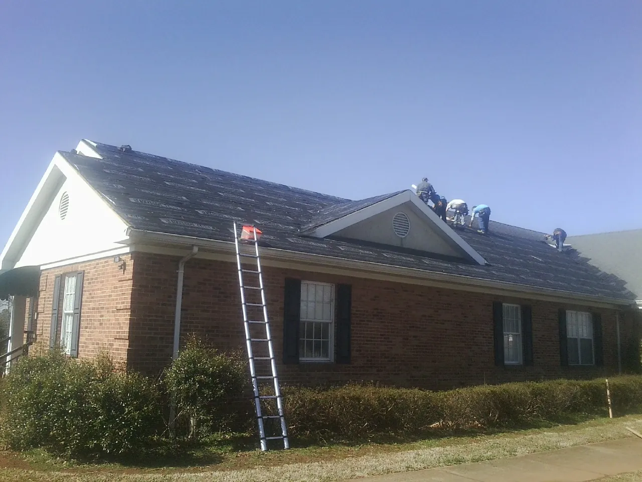 Crew tearing shingles off a brick ranch roof with a ladder set — Metal Roof Repair work by our crew in Marne